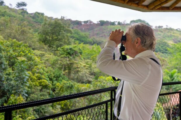 Tony Juniper looks out over a balcony at the green tree canopy and wildlife beyond through his binoculars