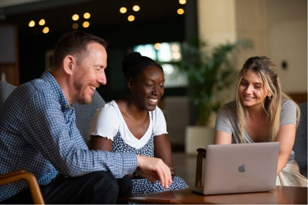 Three people looking at a laptop screen in an office, smiling and talking.