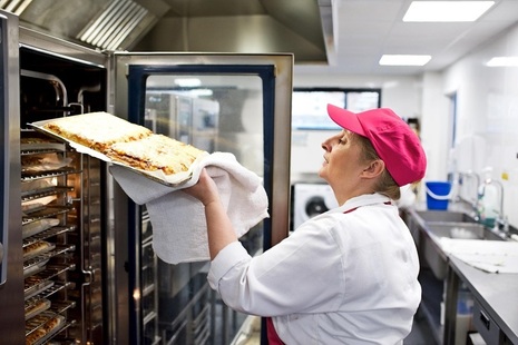 A person in a professional kitchen, wearing catering clothing, removing a tray of hot food from an industrial oven.