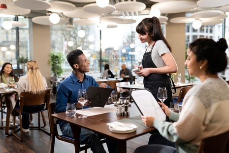 Two people at a small table in a brightly lit restaurant ordering food from a server.