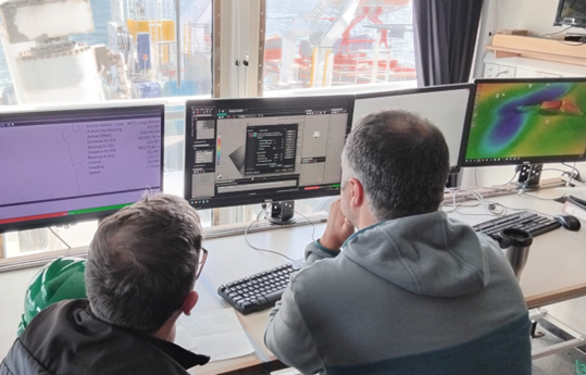 Two people in front of computer screens onboard a vessel reviewing seabed mapping data.