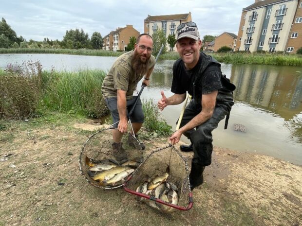 Two Environment Agency officers smile at the camera, each with a long-handled net filled with fish.