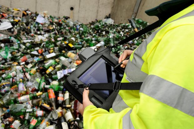 A person in a hi-vis jacket inputs info into a tablet computer whilst looking at waste glass bottles.