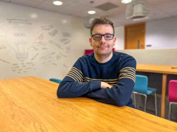 Man in glasses in office setting sat with arms crossed and smiling to camera.