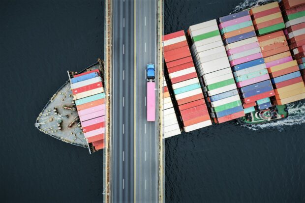 Aerial view of a container ship passing beneath a suspension bridge. Semi truck with pink cargo container crosses above.