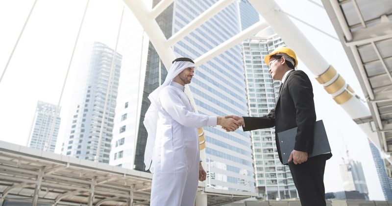 Man in a suit and hard hat shaking hands with a man dressed in white, with a modern city in the background