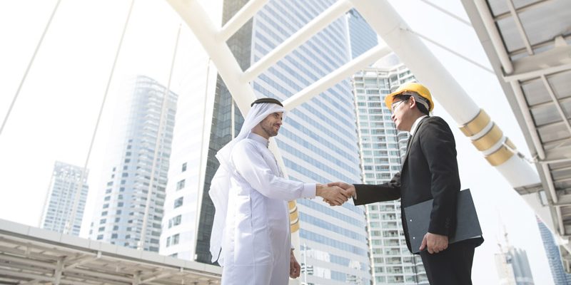 Man in a suit and hard hat shaking hands with a man dressed in white, with a modern city in the background