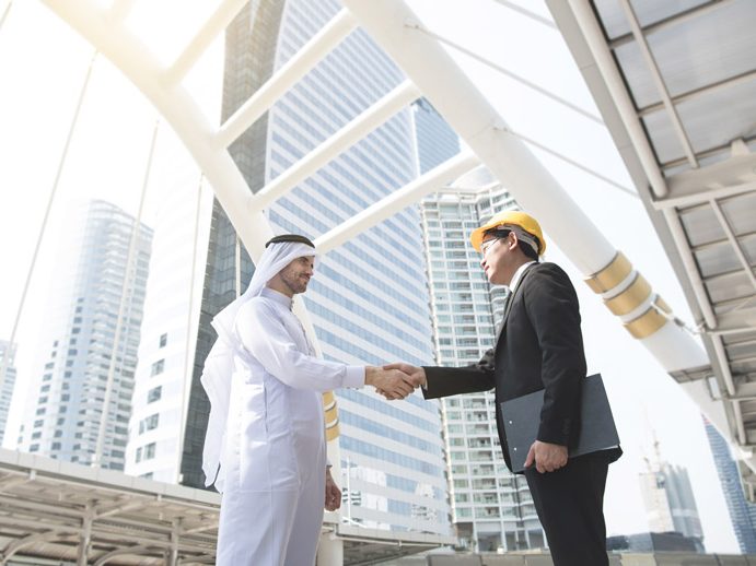 Man in a suit and hard hat shaking hands with a man dressed in white, with a modern city in the background