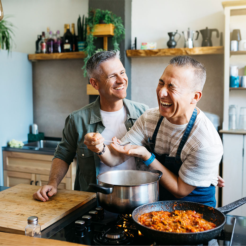 Photograph of two men cooking together in a kitchen, with one man wearing an apron stirring a pot on the hob and the other standing close behind him. Kitchen setting includes wooden shelves with plants and bottles, a frying pan with cooked food on the hob, and a wooden chopping board on the worktop.