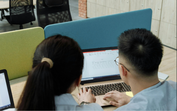 A man and a woman in an office using a laptop. The picture shows the backs of their heads.