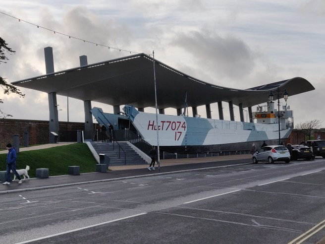 Broadside view of LCT 7074 in preservatiion at the museum, painted in camouflage battleship grey colours, set under an exterior canopy with steps leading up to her open bow doors.