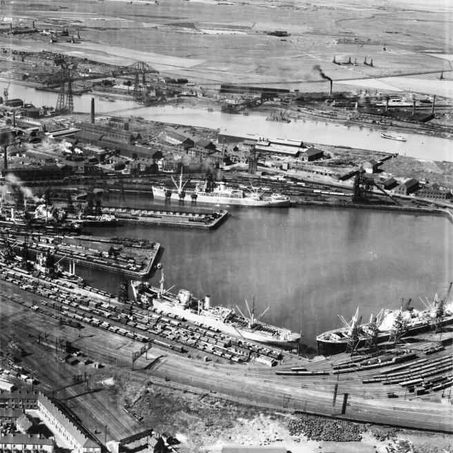 Historic B&W aerial photo of rail lines surrounding a dock in the foreground, with several ships in it. In the middle ground is the Tees with the Transporter Bridge to left, and in the background a rural landscape contrasting with the industrial riverscape