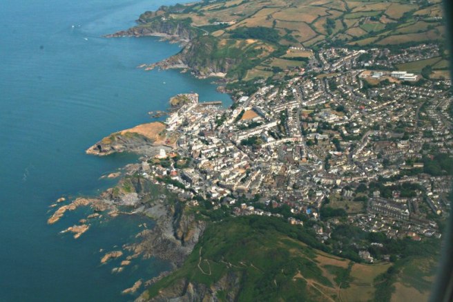 Aerial view of a coastal town with the sea to the left of image, showcasing a mixture of residential buildings, hills, and rocky shoreline with agricultural land in the background.