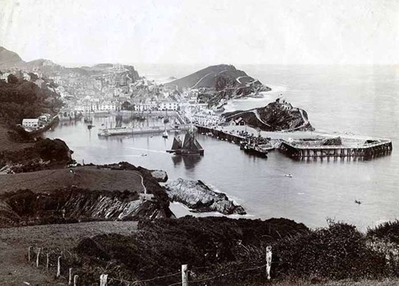Historical black and white photograph of a coastal harbour looking down from the top of the cliff to the water below, centred on a sailing ship leaving harbour. Beyond the harbour in the background can be seen encircling hills and cliffs sheltering the harbour from the open sea beyond. 