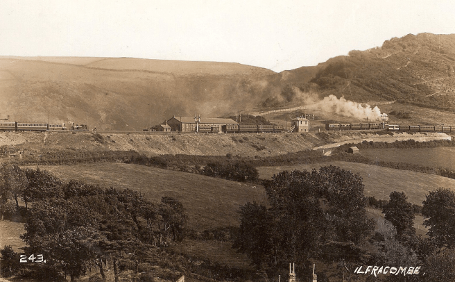 Historic sepia photograph showing a train travelling along to right with a noticeable head of steam, with a station and buildings in centre background, set in a rural landscape of rolling hills beyond. 