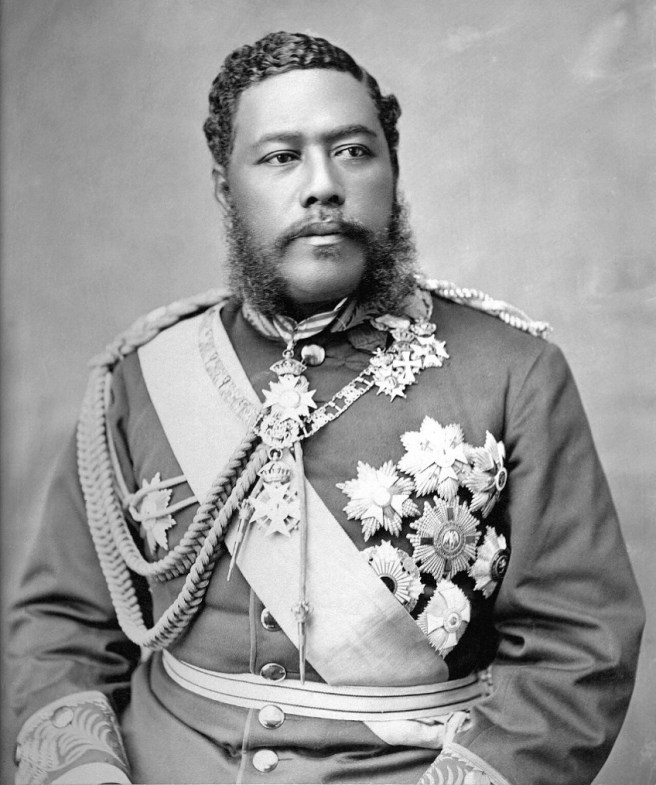 Historic black & white portrait photo of indigenous Hawaiian with black curly hair and a curly beard and moustache in a European-style braided military uniform covered with extensive regalia