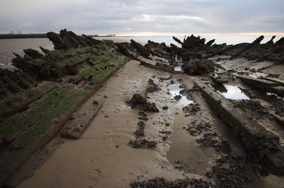 Modern digital colour photograph of the 'ship shape' of the exposed hull timbers full of sand and pools of water, looking out towards the sea. 