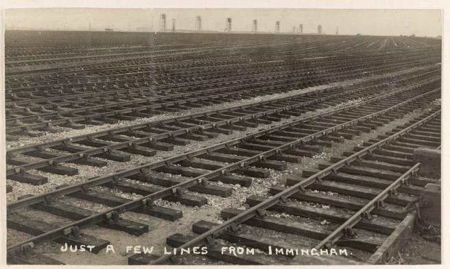 Historic sepia postcard of parallel rows of multiple railway lines stretching into the distance, with the vertical posts of the coal hoists of the dock just visible to the top of the photograph. The postcard bears the punning title on the front 'Just a few lines from Immingham'