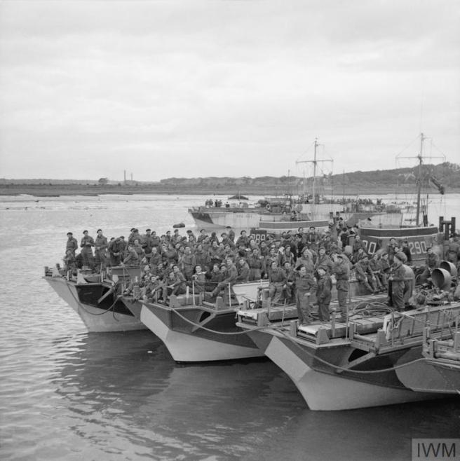 Historic black and white photograph of several landing craft roped together in harbour, with men in military uniform gathered on their decks