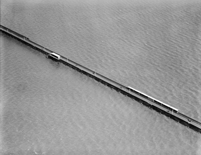 Historic black and white aerial view of the Southend Pier Railway stretching over water from top left to lower right: the railway is seen in operation with a train towards lower right