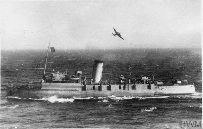 A vintage black and white photograph of the ship in wartime livery, with an aeroplane flying overhead.