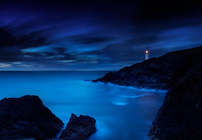 Simple modern colour digital photograph of the land silhouetted in black to foreground (rocks) and right (cliffs) of image, with the white lighthouse and its light appearing to right background. The blue sea comes in from the left background to right foreground, and dark clouds are visible in the dark blue sky.