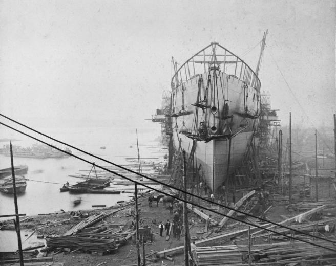 Historic black and white photograph of ship under construction on the bank of the Thames on the right, with barges on the river to the left, seen from behind scaffolding in the foreground