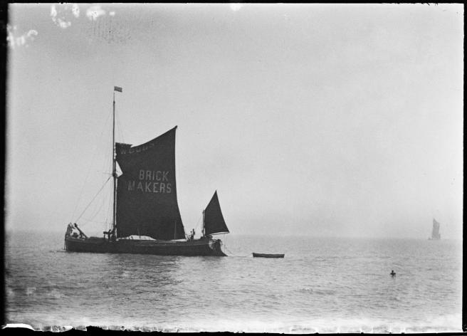 Historic black & white photograph of a Thames barge under sail, to left foreground, towing a boat. To the right another Thames barge looms out of the mist in the distance.