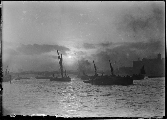 Historic black and white photograph of barges with furled sails in centre ground on a moonlit night.