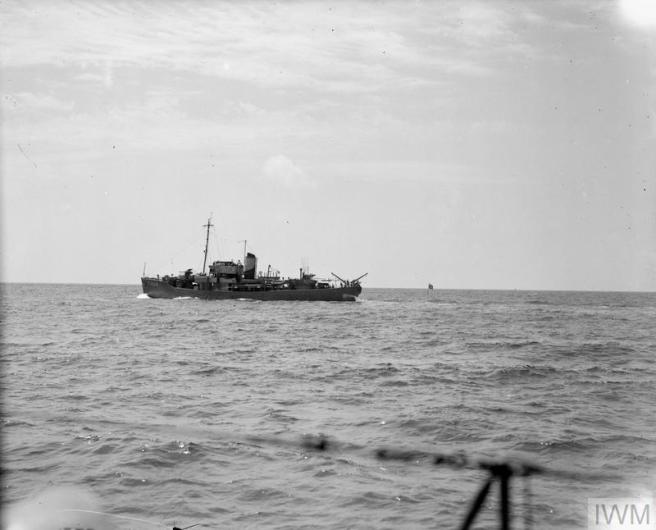 Historic B&W photo of ship seen in port profile view at sea from the railings of another ship in the foreground. Behind her a marker rising out of the sea can be seen (image can be explored in more detail by following the link in the caption)