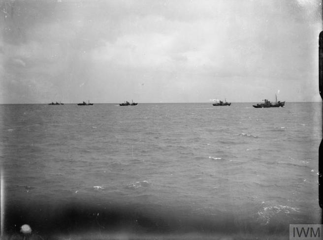 Historic black & white photo from the railings of a ship at sea which blur the bottom foreground, looking towards five minesweepers at work on the horizon