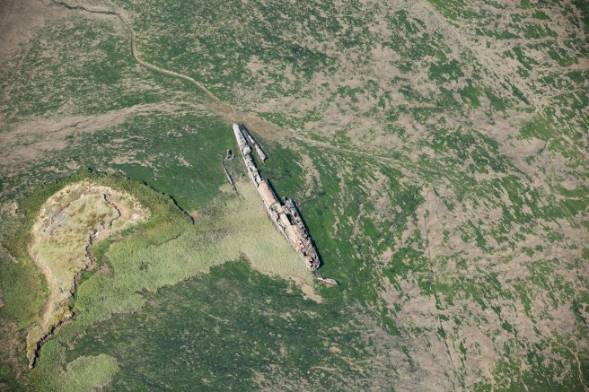 Aerial photograph of green saltmarsh with remains of submarine hull in centre, orientated NNW-SSE, the outline of the hull being broken at the upper right.