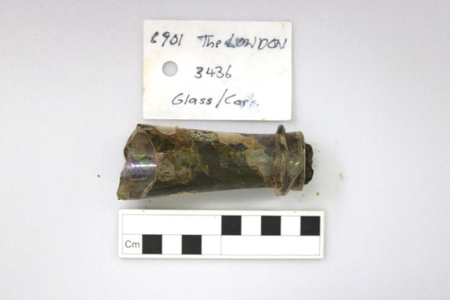 Archaeological record photo of glass bottle neck with scale and tag.