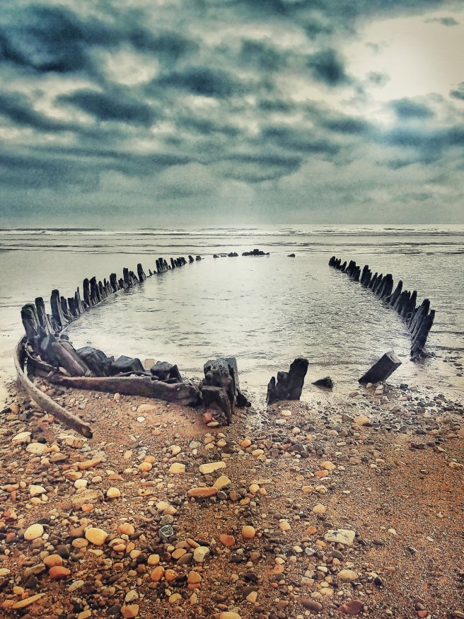 The timbers of a wreck on a shingle beach fill up with water as the tide comes in, under heavy cloud cover.