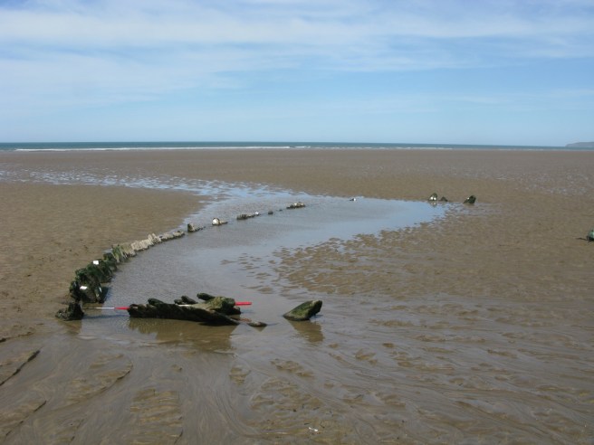 Stretch of sandy beach with a strip of water in the middle of the photograph, surrounding the timbers of a wreck protruding from the sand, set against a wide blue sky.