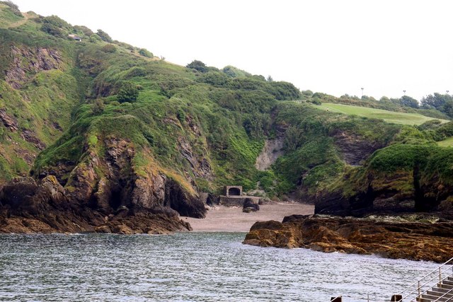 Photograph of rocky cove with steep, hilly sides covered in green. The tide is nearly at the entrance to the cove, which is very narrow.