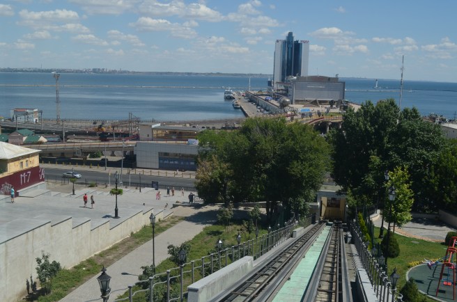 View of port city, with tree-lined plaza in foreground, looking out to sea over port infrastructure, and a blue sky beyond.