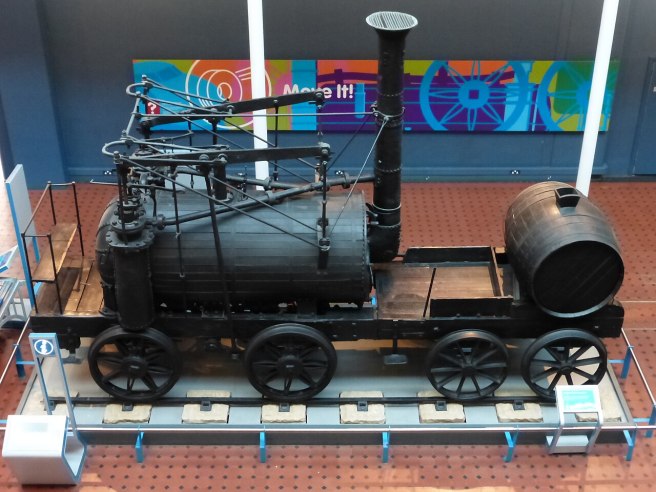 View of Wylam Dilly locomative from above in a museum setting, giving prominent attention to its tall chimney and small size