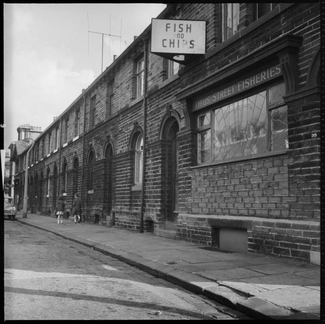 Streetscape of brick terraced houses with round-headed door frames and windows. To right is a shop window bearing a stained glass Art Deco sunrise design with the legend 'Titus Street Fisheries' at the top.