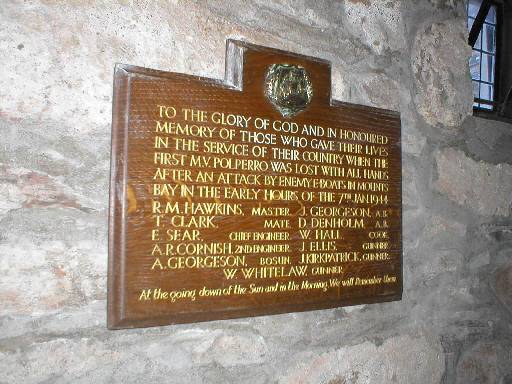 Modern colour photograph of wooden war memorial plaque with text picked out in gold, naming the men of the MV Polperro and the date of loss, mounted on the roughcast wall of a rural church.