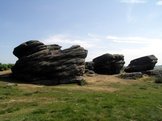 Modern colour photograph of three rock formations tilted upwards as if they were ships ploughing through seas, instead of the grass platform on which they sit, seen against a blue sky.