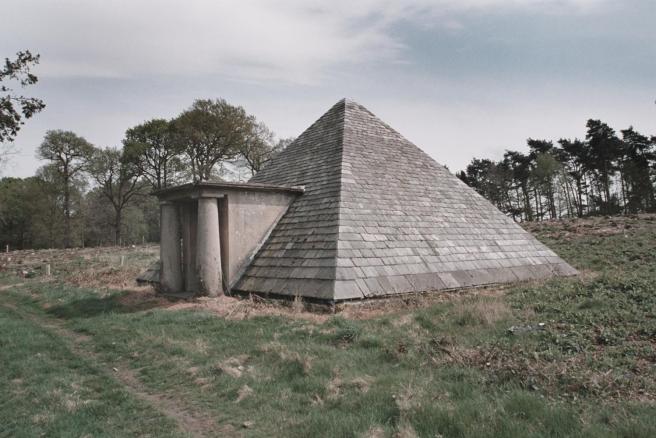 Modern colour photograph of stone pyramid with entrance porch flanked by classical columns, seen against a backdrop of tall trees on a cloudy day.