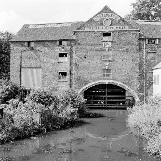 Historic B&W photograph showing the corn mill seen from the canal towards the boat arch, with the text 'F E Stevens Ltd./Trent Corn Mills in the gable of the mill, with a plaque saying 'No.2 Mill' below a window.