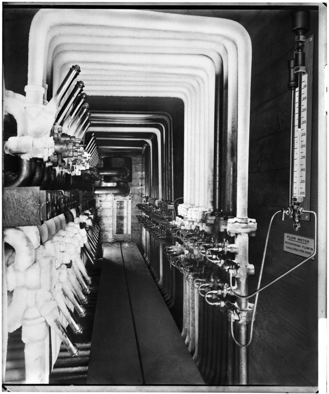 Historic B&W photograph of a long row of pipes with a flow meter connected to feeder valves on the right. The pipes are angled upwards, across the ceiling, and run down again opposite, with two rows of valves.
