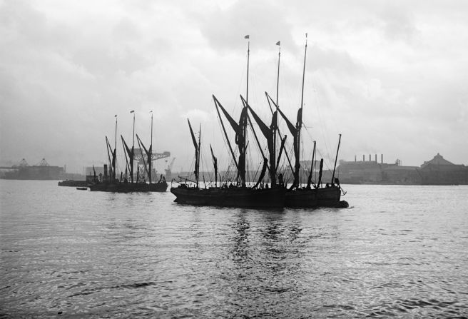 Two groups of Thames barges moored on the river, with their sails furled. Behind them lies an industrial landscape of chimneys and dock cranes on the far bank of the river.