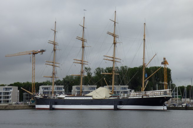 Modern colour photograph of four-masted museum ship with four bare masts and a crane beyond at left, against a grey sky.