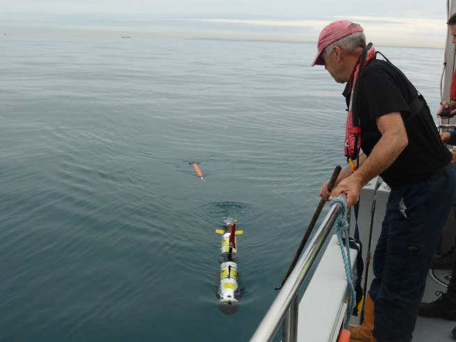 Man dressed in black to the right of the image looks over the railing of a boat at an underwater vehicle on the surface of the water to his left.