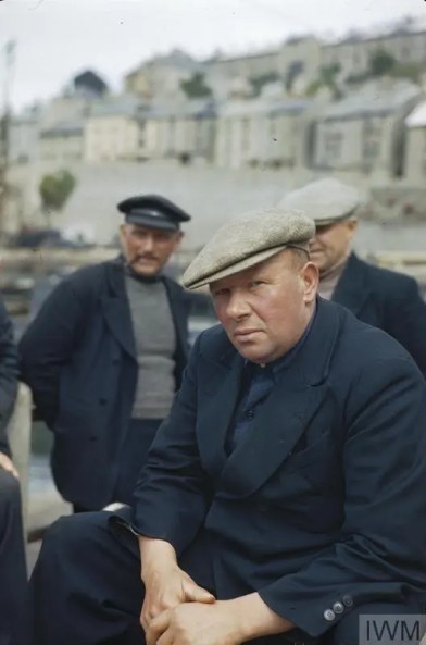 Historical colour photo of a group of three fishermen in typical navy blue fishing gear with caps on their heads, with a blurred view of terraces behind them.