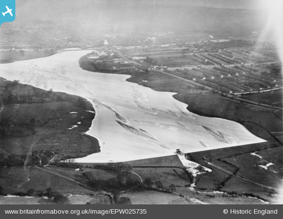 Historic black & white photograph of a frozen lake seen from the air, surrounded by fields and a housing estate to the upper right of the photo.