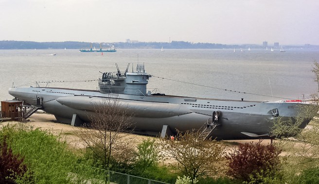 Modern colour photograph of a propped submarine with visitor access steps, sitting on a beach, with passing vessels in the background. In the foreground are trees.